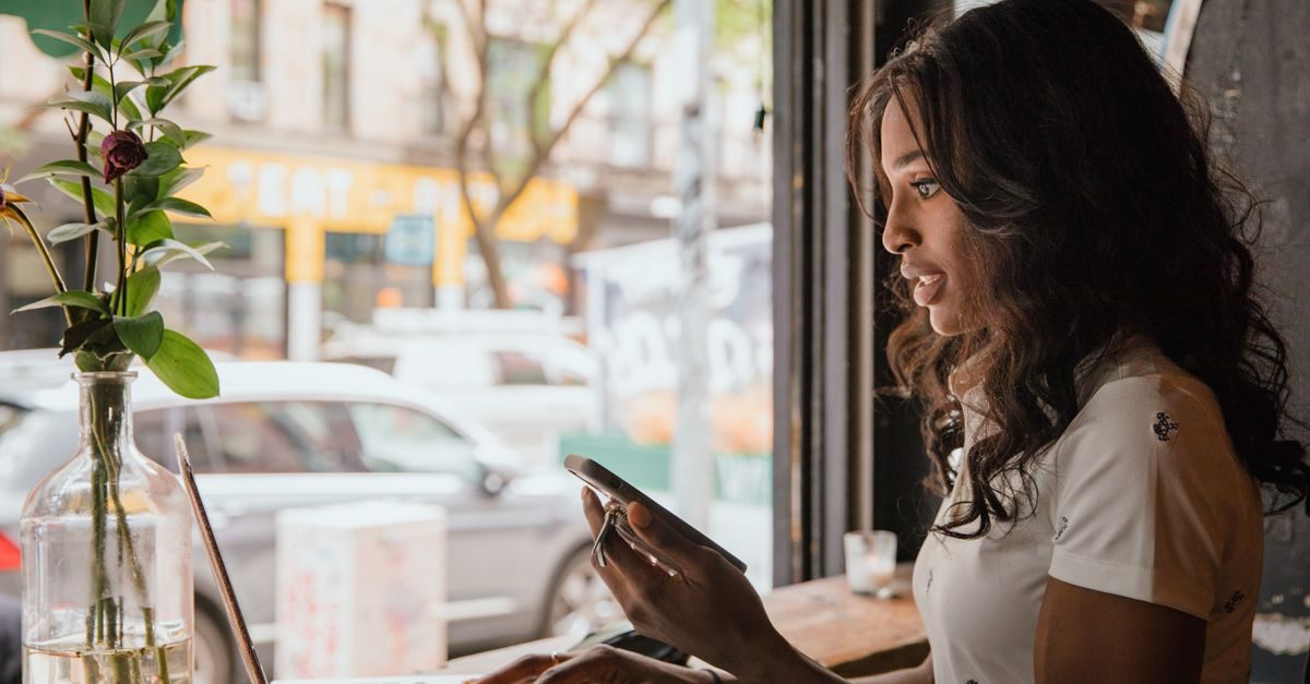 Jonge vrouw met lang donker haar kijkt geconcentreerd naar haar smartphone in een sfeervol café met uitzicht op straat