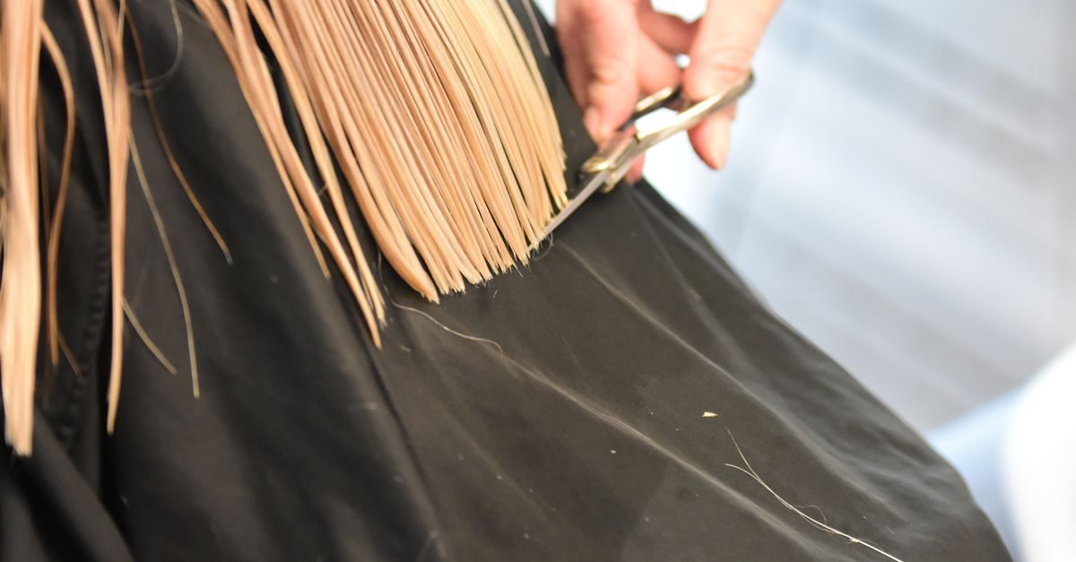 Friseur schneidet mit einer Schere die Spitzen von langem blondem Haar, das durch einen schwarzen Umhang geschützt ist