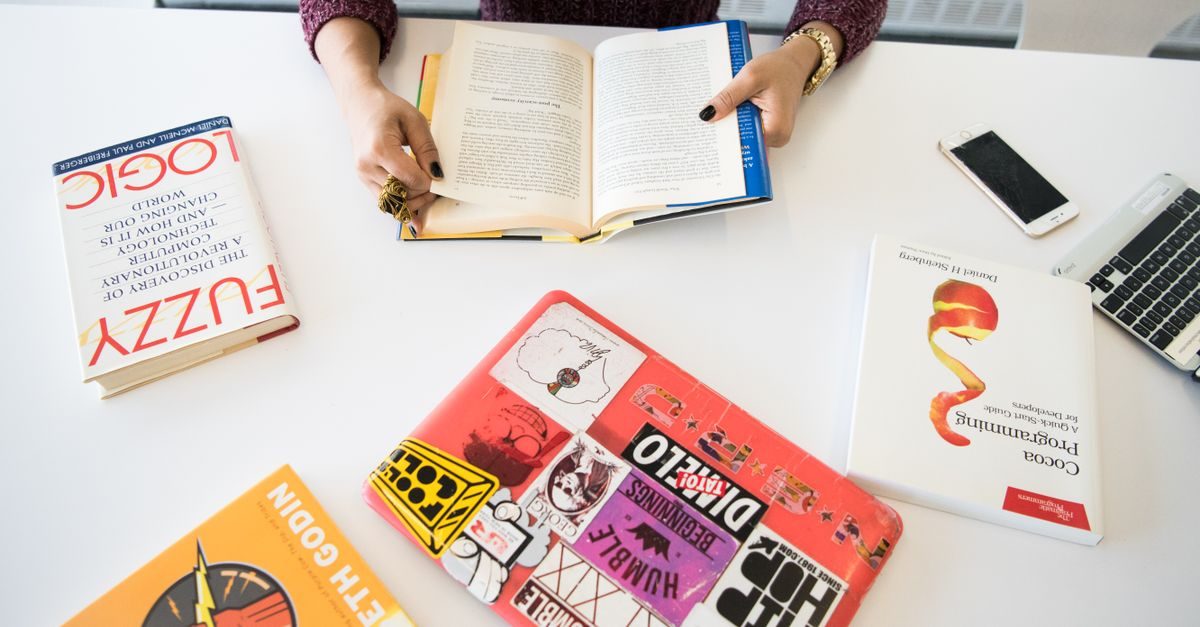 Person liest an einem weißen Schreibtisch mit Fachbüchern, einem Laptop und einem Smartphone in konzentrierter Atmosphäre