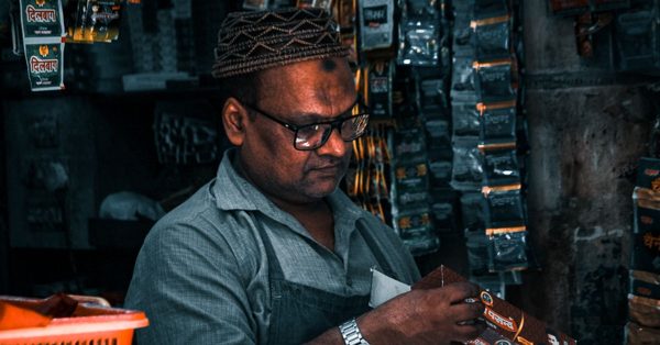 Hombre con gafas y gorro tradicional trabajando concentrado en su pequeña tienda llena de productos colgados