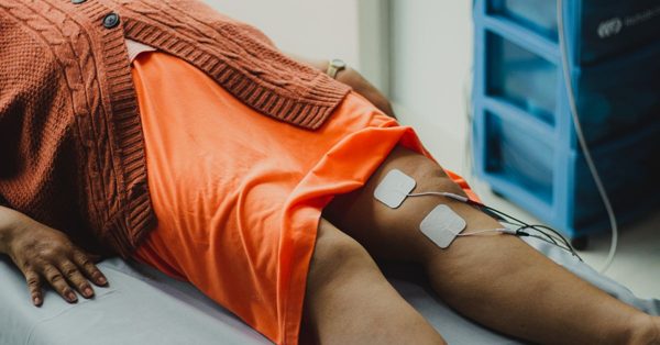 A person lying on a clinic table receives electrical stimulation therapy on their knee with two adhesive electrode pads