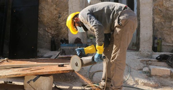 A worker in a yellow hard hat and protective gloves uses an angle grinder to cut a metal sheet at a construction site