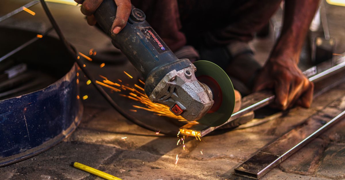 A worker uses an angle grinder to cut a metal bar, creating a shower of bright orange sparks in a workshop setting