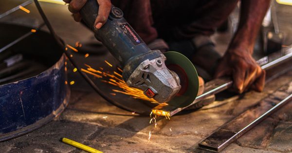 A worker uses an angle grinder to cut a metal bar, creating a shower of bright orange sparks in a workshop setting