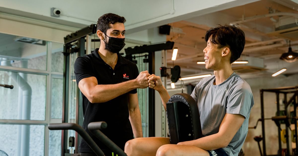 A masked fitness trainer and a smiling client share a fist bump in a modern gym setting