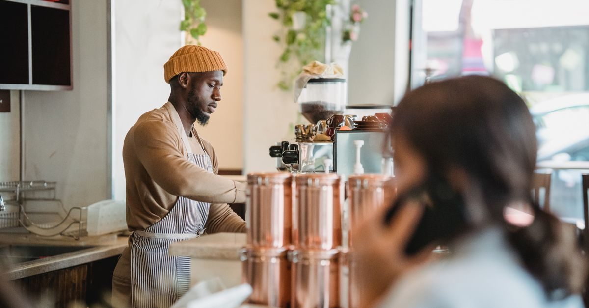 Ein konzentrierter Barista in gelber Mütze und Schürze bereitet hinter einer Kaffeetheke ein Getränk zu