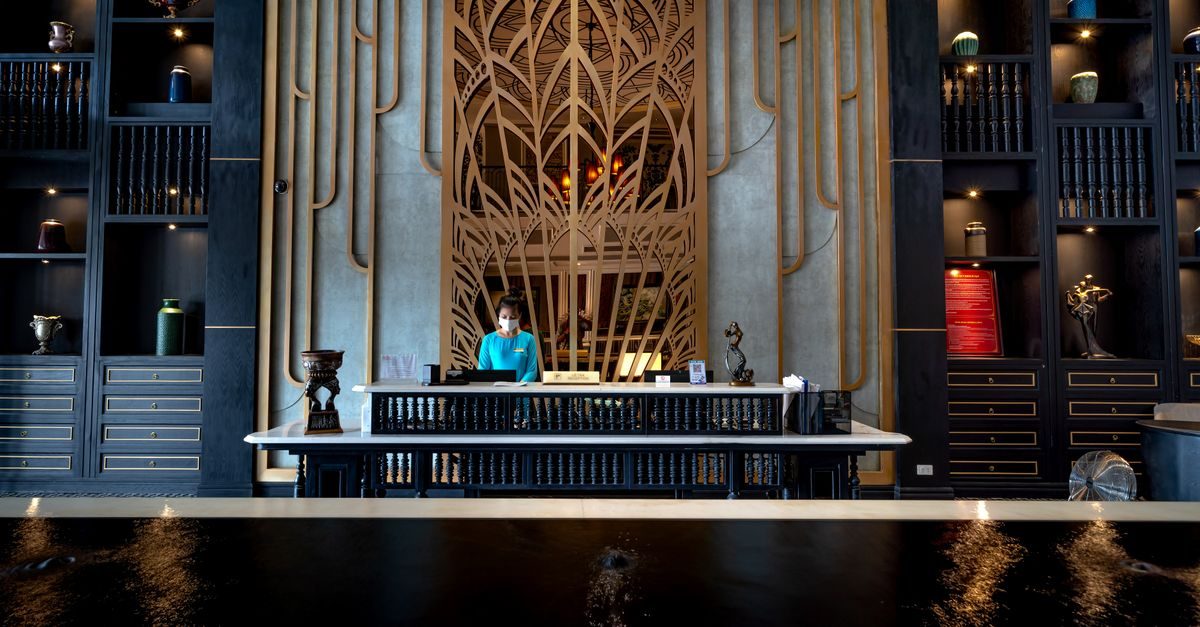 Recepcionista con mascarilla tras un elegante mostrador de hotel con estanterías decorativas y fondo geométrico dorado