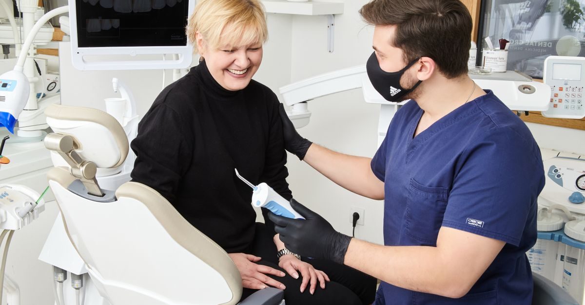Ein Zahnarzt mit Mundschutz berät eine lächelnde Patientin in einer modernen Zahnarztpraxis mit einem Mundduschen-Handstück