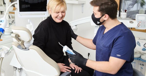 Ein Zahnarzt mit Mundschutz berät eine lächelnde Patientin in einer modernen Zahnarztpraxis mit einem Mundduschen-Handstück