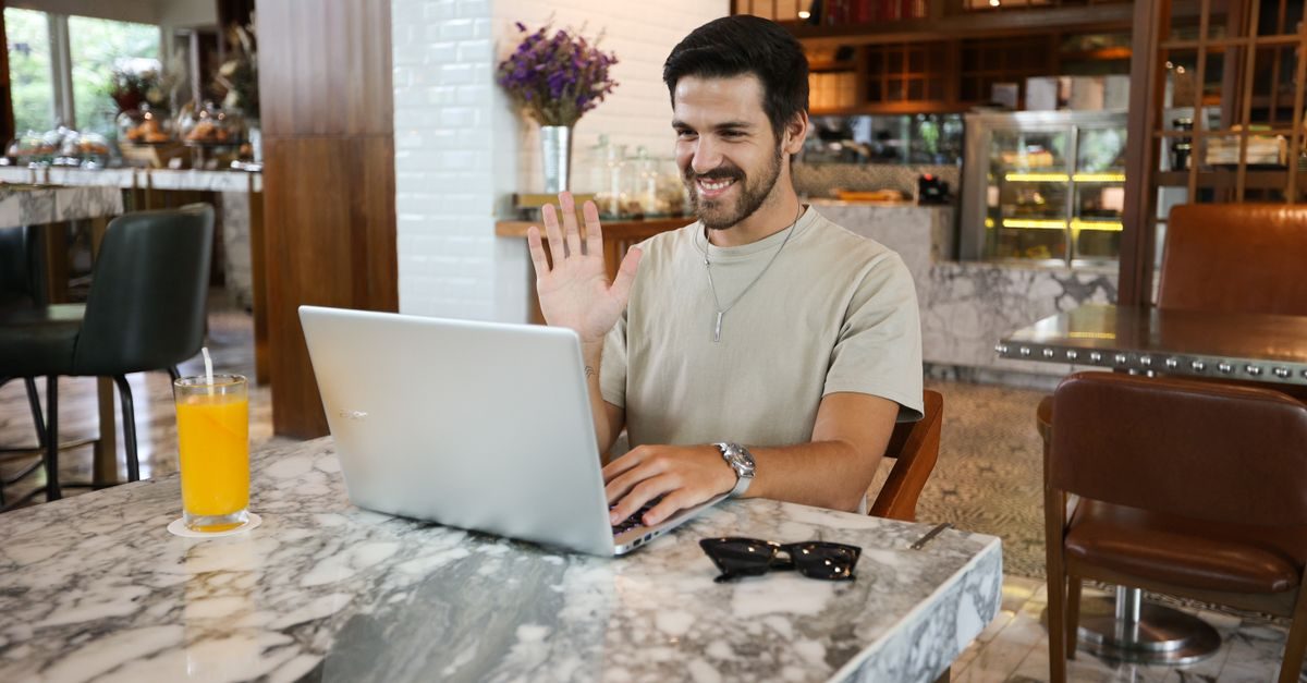Glimlachende man zwaait naar zijn laptop tijdens een videogesprek aan een marmeren tafel in een sfeervol café