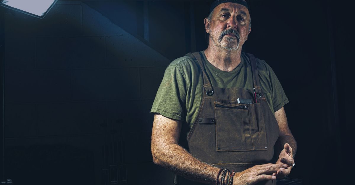 Ervaren vakman met snor en werkschort kijkt serieus in een donkere werkplaats met sfeervolle belichting