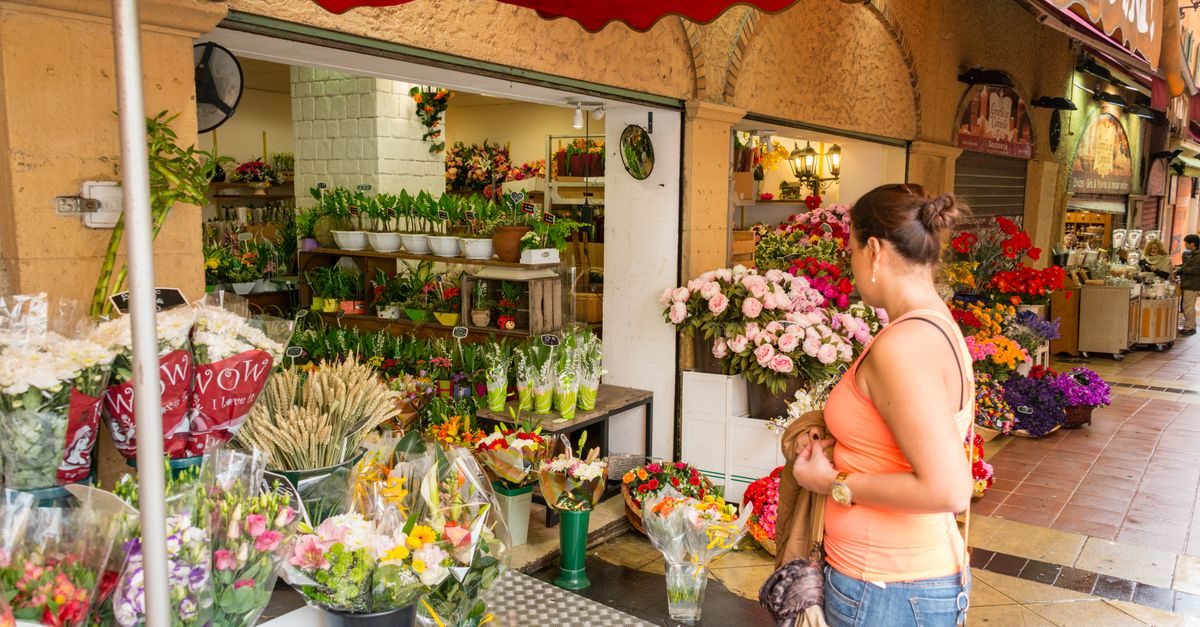 A woman in an orange tank top browses a vibrant street-level flower shop filled with bouquets and potted plants