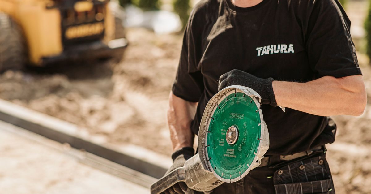 A worker in a black uniform holds a circular saw with a green blade at a construction site with a yellow machine behind