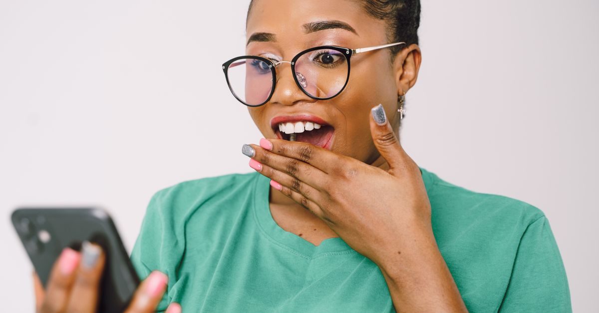 A surprised woman in a green shirt and glasses looks at her smartphone with her hand over her open mouth