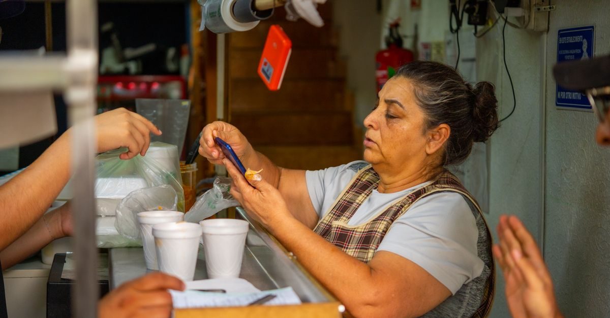 Una donna al bancone di un locale guarda lo schermo di uno smartphone mentre serve dei clienti in un ambiente informale
