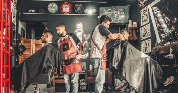 Två frisörer klipper kunder i en barberarsalong med väggdekorationer och en röd telefonkiosk i bakgrunden
