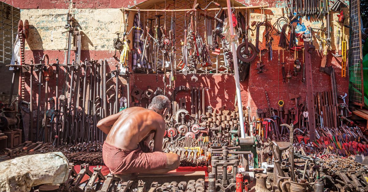 A shirtless man sits in front of a wall covered with a vast collection of vintage metal tools and hardware in a sunny area