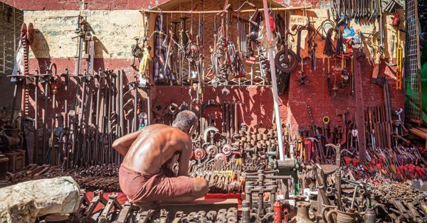 A shirtless man sits in front of a wall covered with a vast collection of vintage metal tools and hardware in a sunny area