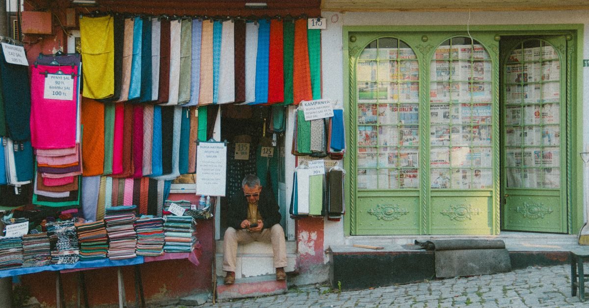 Un homme assis devant une boutique de tissus colorés aux vitrines recouvertes de journaux dans une rue pavée