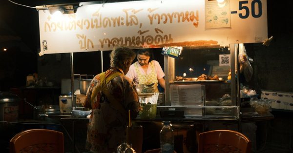 Een vrouw bereidt eten in een verlichte Thaise straatkraam in de avond terwijl een klant toekijkt
