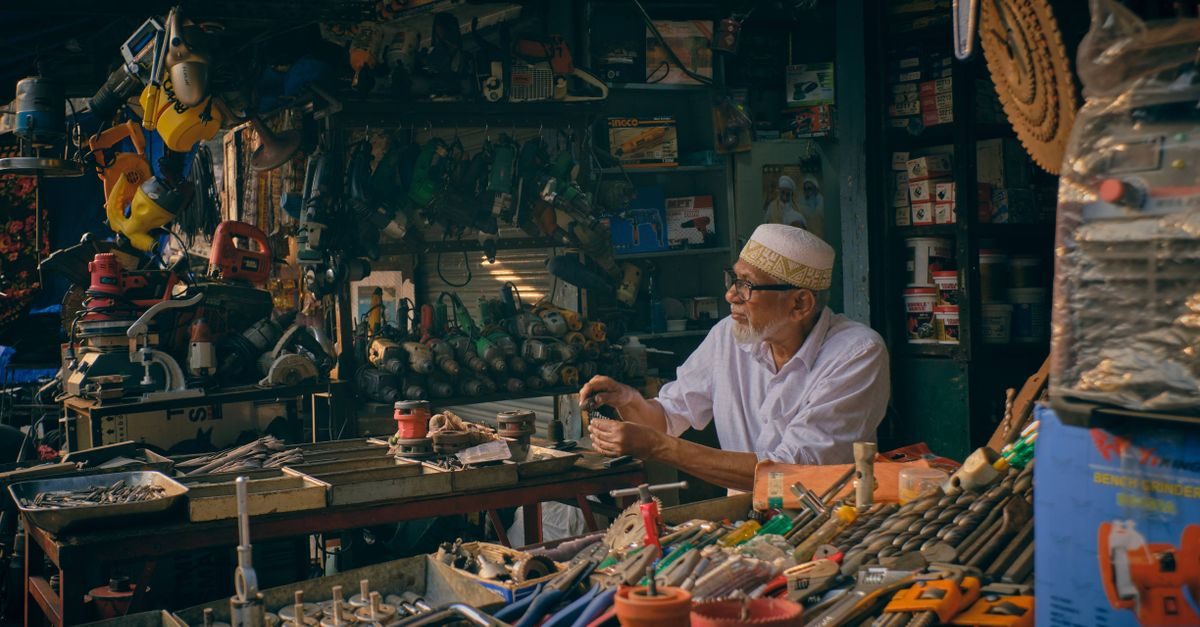 Uomo anziano con copricapo tradizionale lavora concentrato nel suo negozio di utensili edili pieni di attrezzi da lavoro