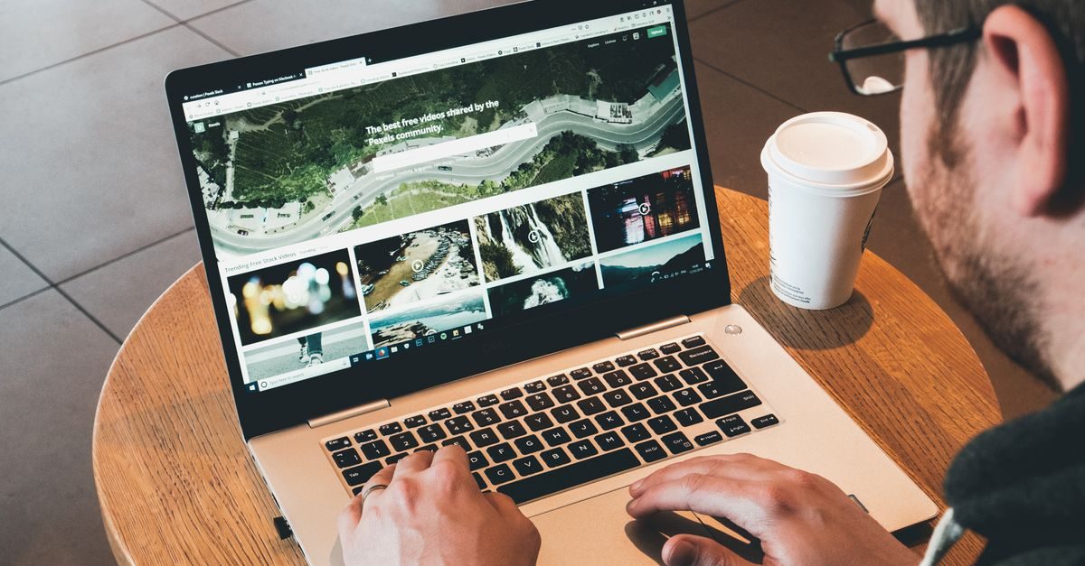 Persoon met bril werkt op een laptop aan een houten tafel met een koffiebeker in een rustige werkomgeving