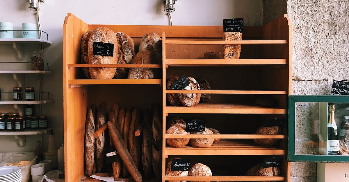 Scaffali in legno di una panetteria con varie forme di pane artigianale, etichette con prezzi e barattoli su mensole laterali