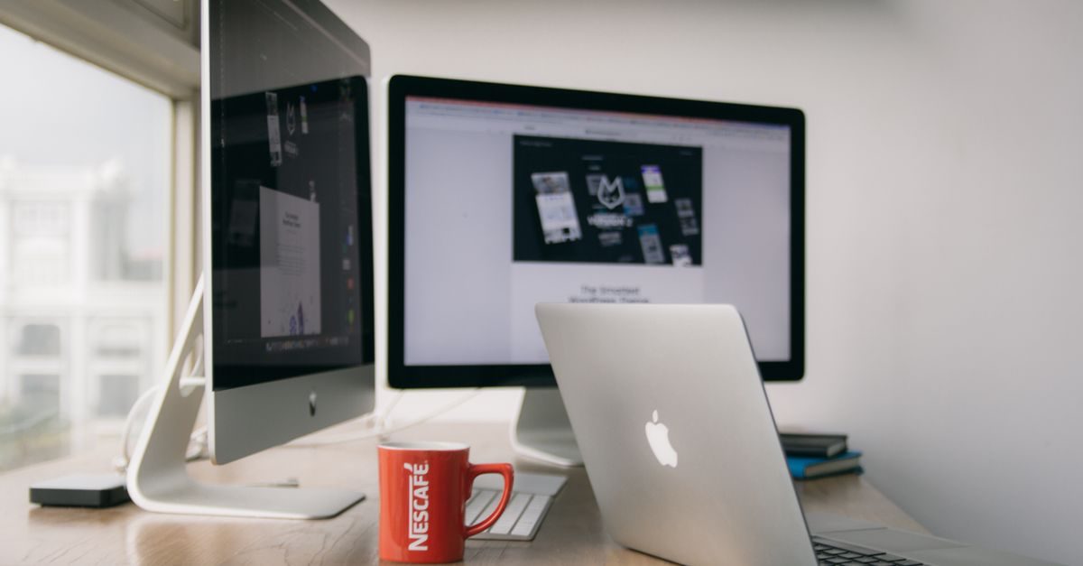 Moderne werkplek met twee computerschermen, een laptop en een rode Nescafé-mok op een houten bureau bij een raam