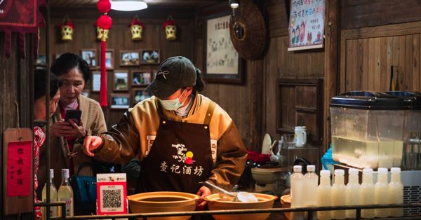 A masked vendor in an apron serves a customer at a rustic wooden stall decorated with red lanterns and framed pictures