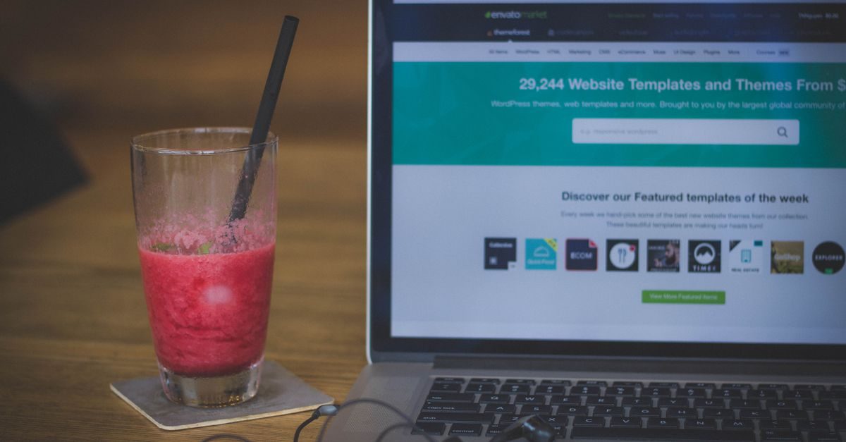 A laptop displaying a website next to a glass of red smoothie with a straw on a wooden table