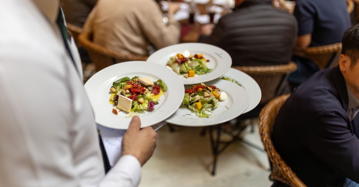 Ober draagt drie borden met salade in een druk restaurant met etende gasten aan tafels