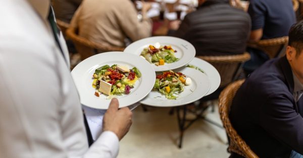 Ober draagt drie borden met salade in een druk restaurant met etende gasten aan tafels