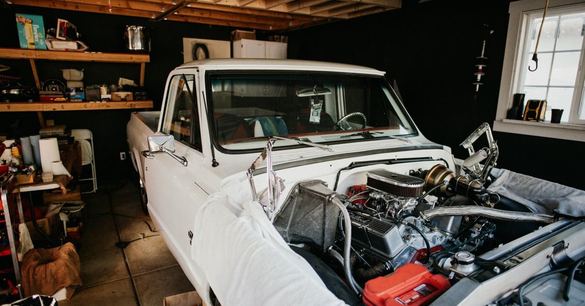 Un pick-up blanc au capot ouvert en cours de réparation dans un garage sombre avec des étagères remplies d'outils