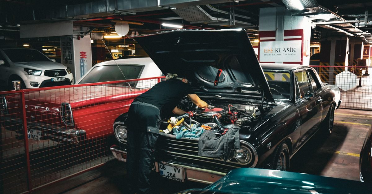 A mechanic works on the engine of a black vintage car in a dimly lit underground parking garage filled with classic vehicles