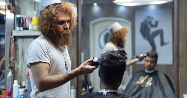 Skäggig frisör med lockigt hår klipper en kund i en barberarsalong med speglar och hårvårdsprodukter i bakgrunden