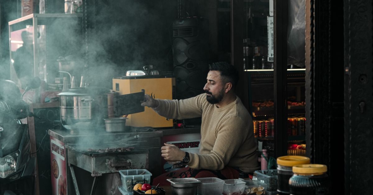 Hombre con barba y suéter beige cocina carne en una parrilla callejera rodeado de humo en un ambiente urbano y nocturno