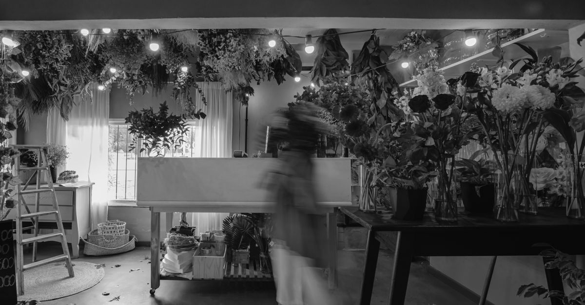 Interno di un fioraio con piante, fiori e luci soffuse, dove una figura umana in movimento crea un effetto sfocato