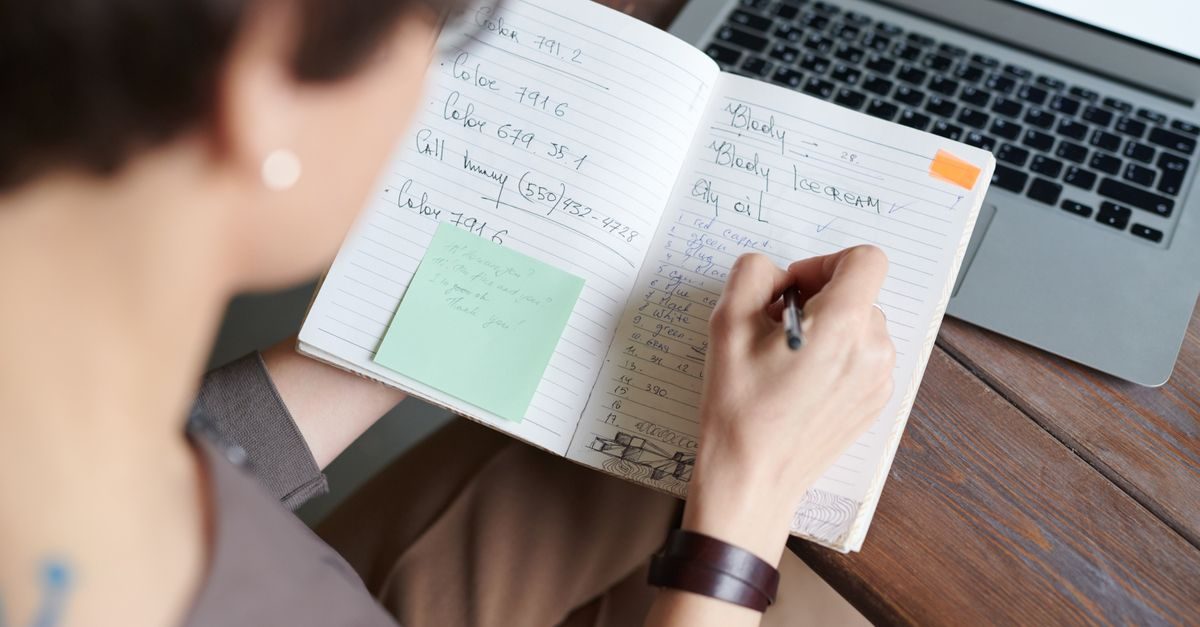 A person sits at a wooden desk writing notes in a journal next to a laptop computer