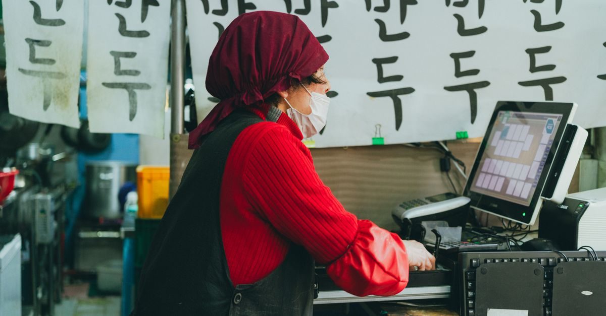 A woman in a red shirt and headscarf wearing a face mask operates a point-of-sale system in a kitchen with Korean signage