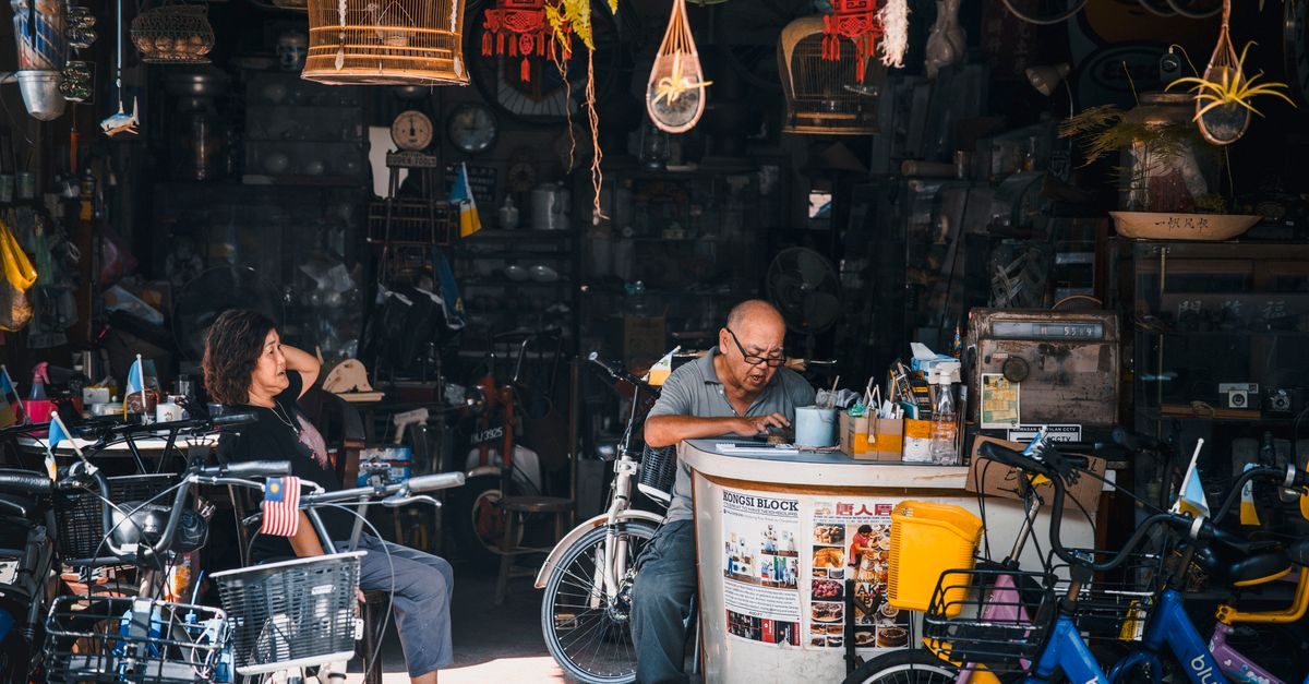 Due persone sedute in un negozio vintage ingombro di biciclette e oggetti antichi, con un uomo che consulta lo smartphone