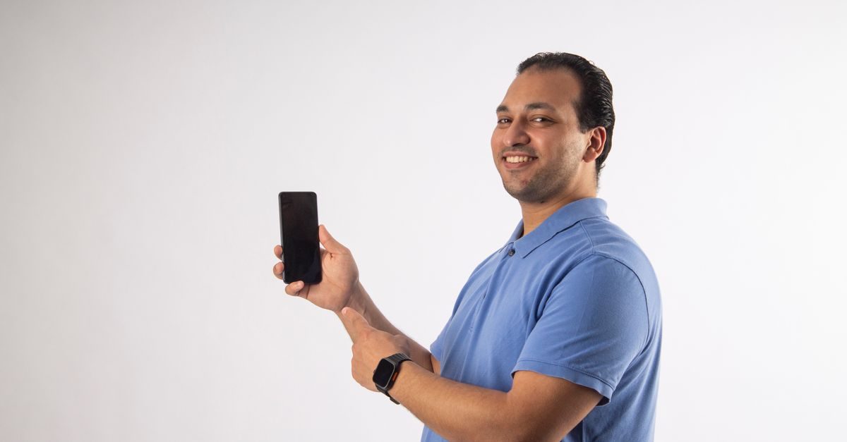 Glimlachende man in blauw poloshirt wijst naar een smartphone die hij in zijn hand houdt tegen een witte achtergrond