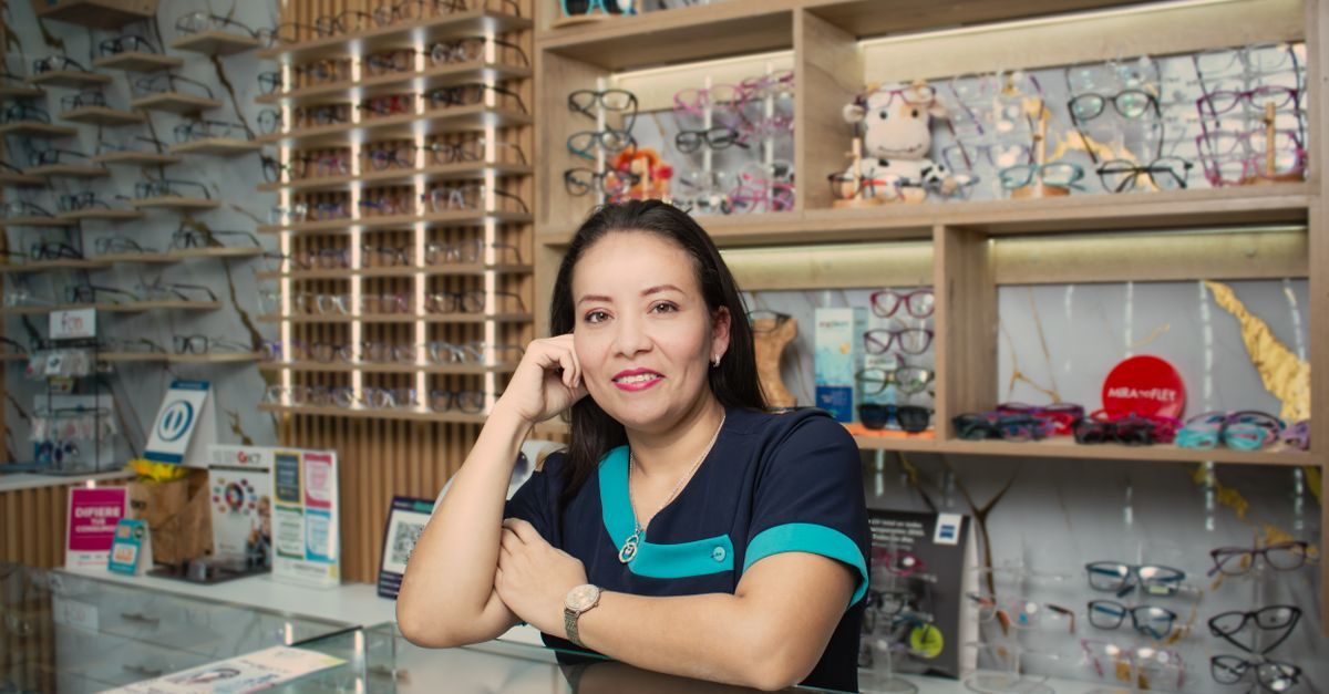 A smiling optician in a blue uniform leans on a counter in a bright store filled with shelves of eyeglasses