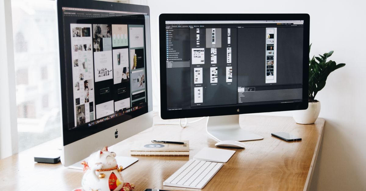 Twee computerschermen met webdesign-ontwerpen op een houten bureau met een gelukskatje en een plant