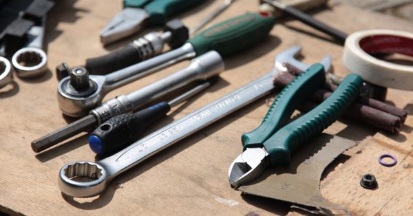 Assorted metal wrenches, pliers, and a screwdriver scattered across a wooden workbench in bright daylight