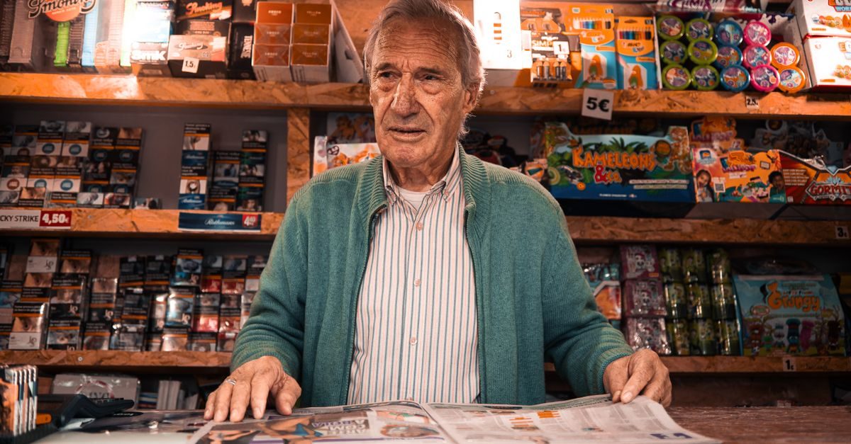 An elderly man in a green cardigan stands behind the counter of a small shop with shelves of goods behind him