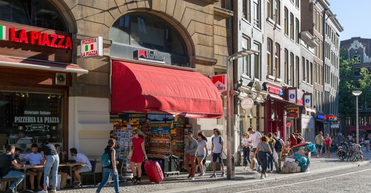Mensen lopen over een zonnige winkelstraat in Amsterdam langs een pizzeria en een tabakswinkel met een rode luifel