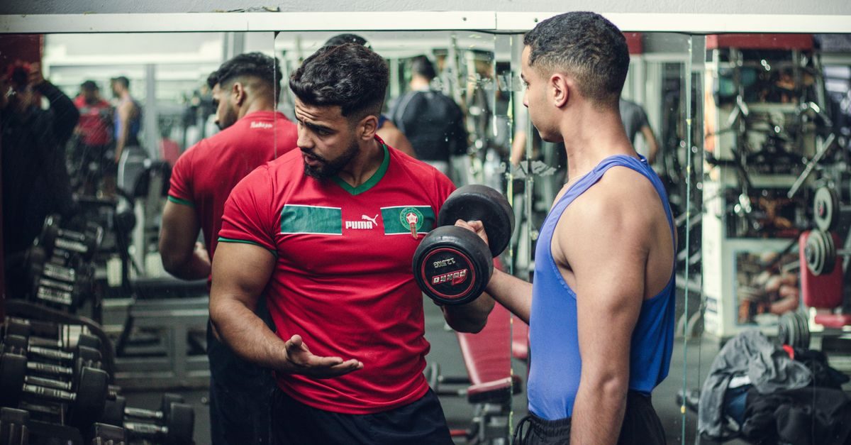 A man in a red Morocco jersey instructs another man lifting a dumbbell in a gym reflected in a large wall mirror