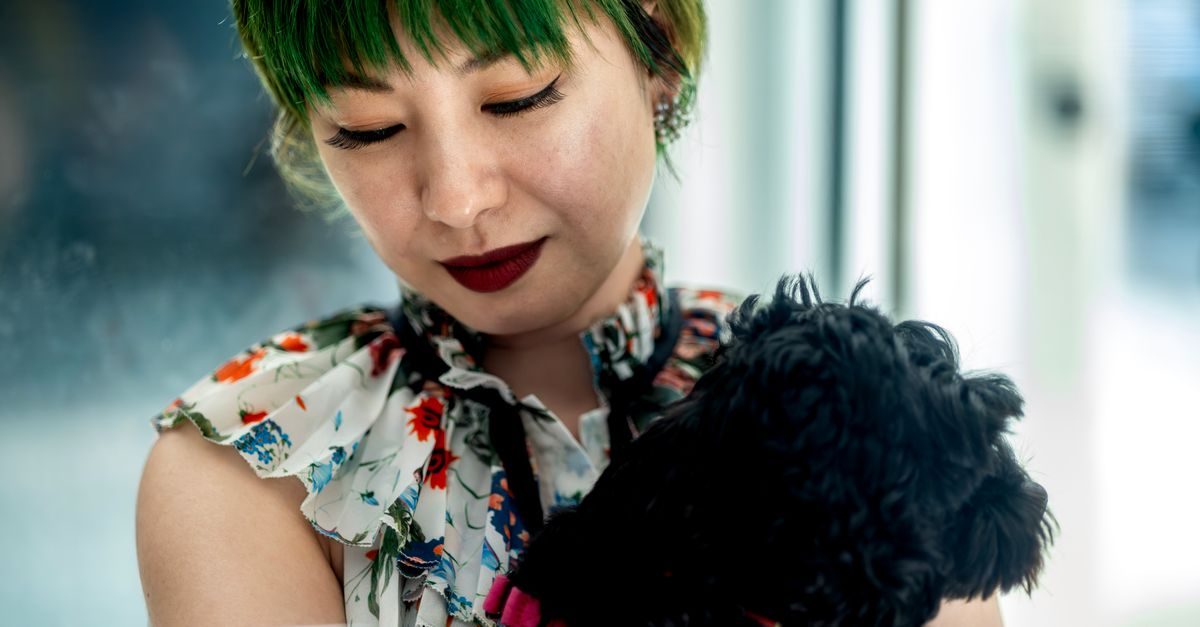 Vrouw met groen haar en bloemenblouse houdt teder een kleine zwarte hond vast in een lichte omgeving