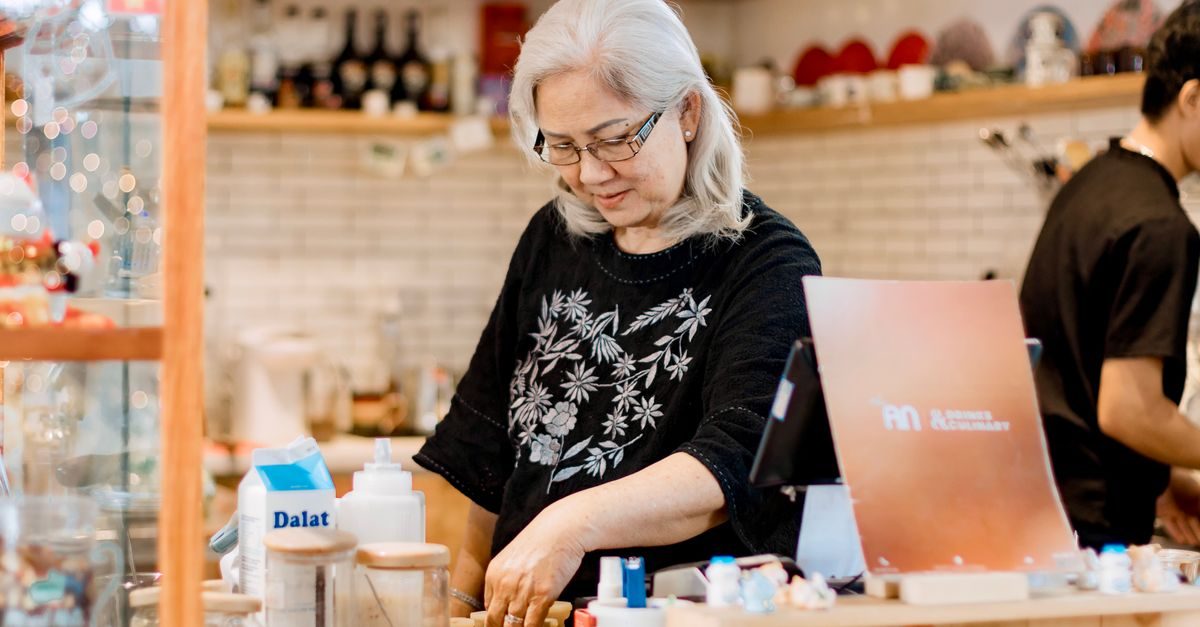 Eine ältere Frau mit Brille arbeitet konzentriert an einer Ladentheke in einem gemütlich eingerichteten Café