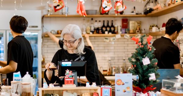 Une femme aux cheveux gris travaille au comptoir d'un café décoré pour Noël entourée de deux collègues